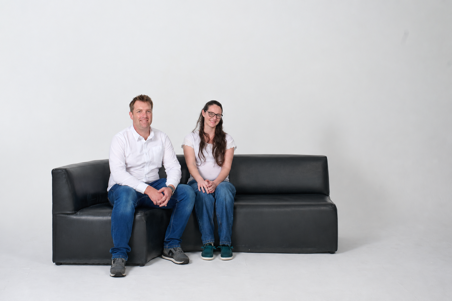 Three people sitting on a black couch against a plain white wall, smiling at the camera, with the man on the left, a woman in the middle, and a man on the right.