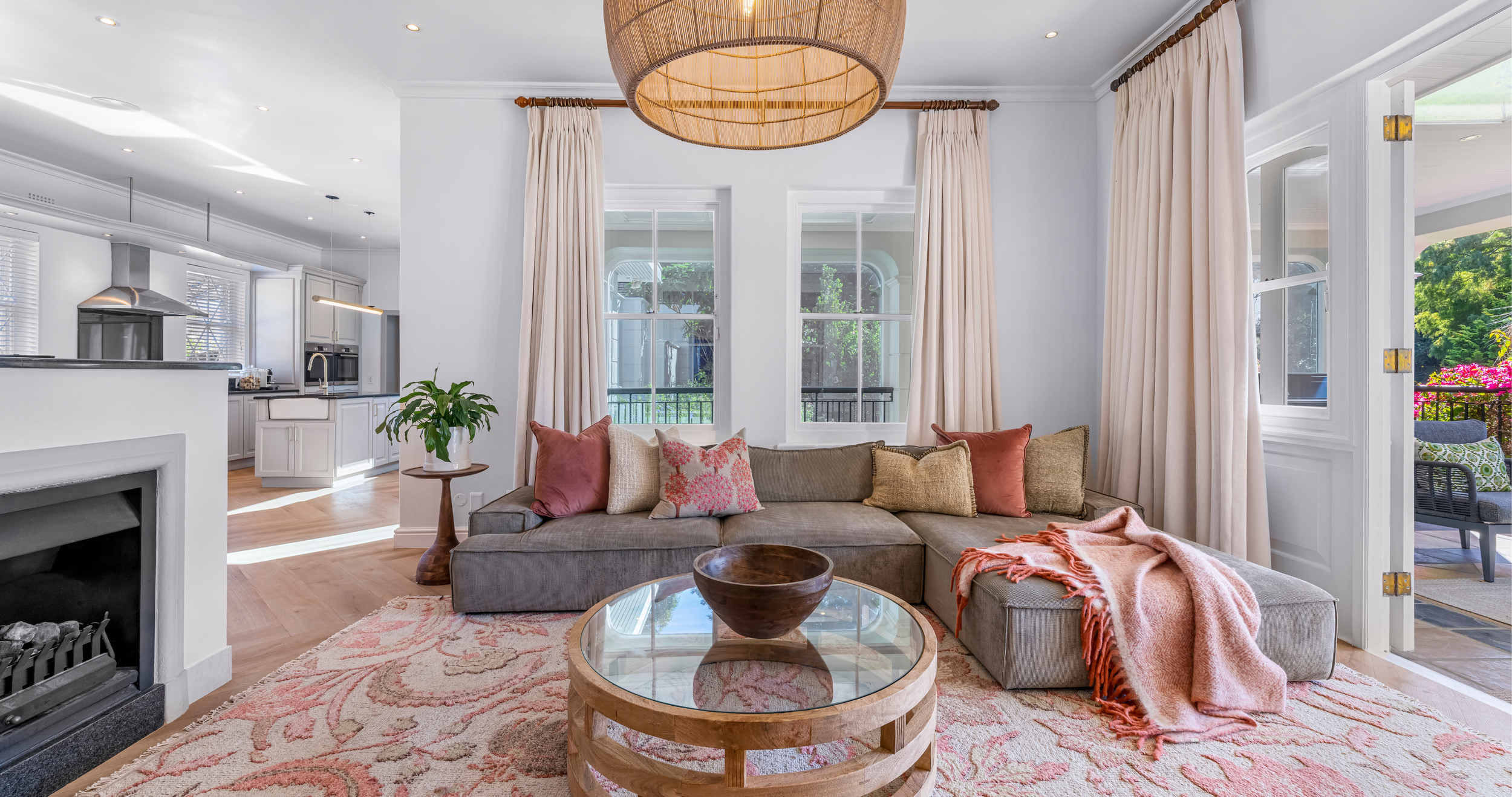 A living room with a beige sectional sofa, pink and beige throw pillows, a pink throw blanket, a round wooden coffee table, and large windows with cream curtains showing a balcony with outdoor seating and greenery.