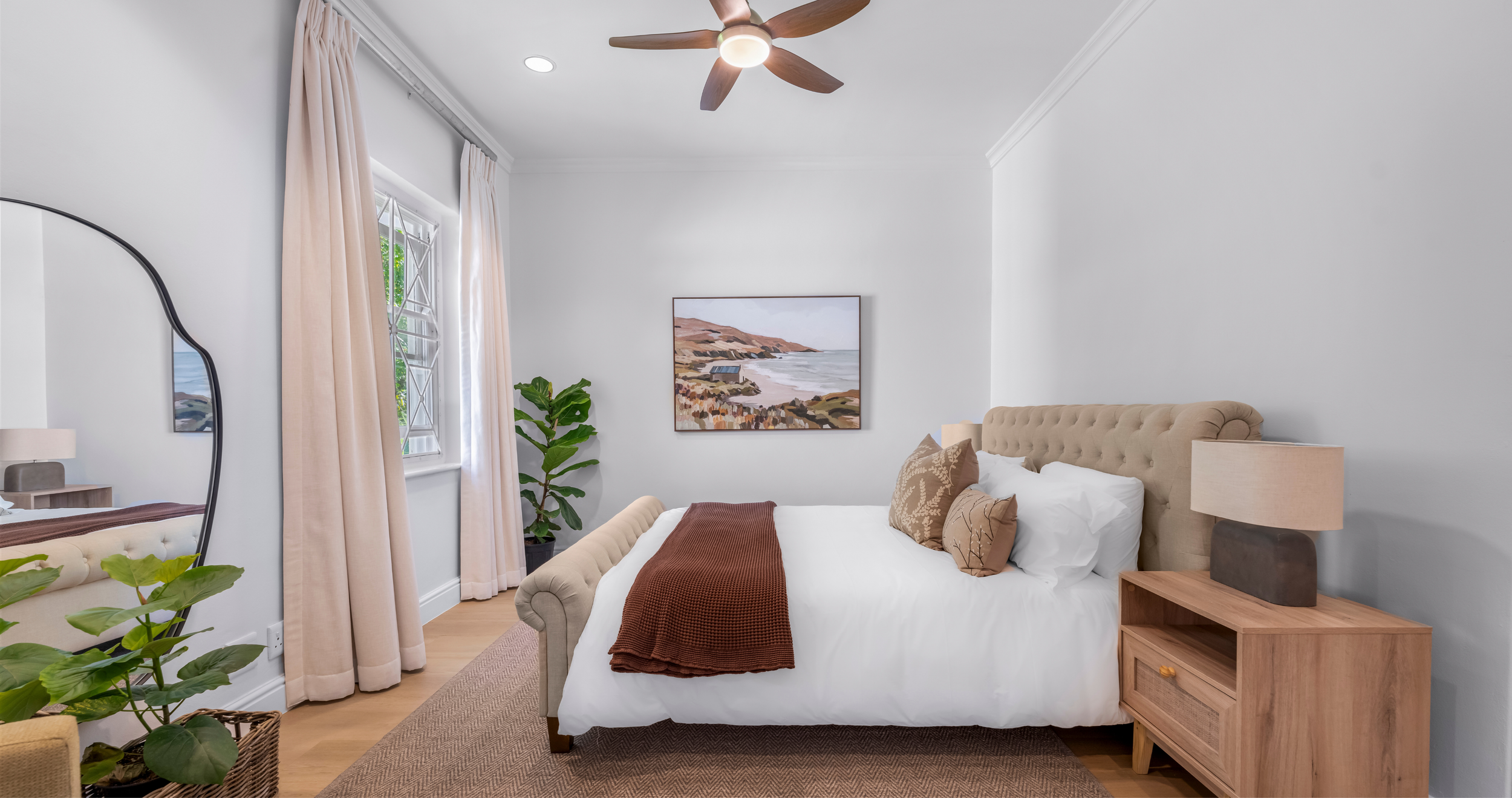 A bedroom with a beige upholstered bed, decorative pillows, and a brown blanket, a wooden nightstand with a lamp, a large indoor plant, a framed landscape painting on the wall, beige curtains, a window, a ceiling fan, and light wood flooring.