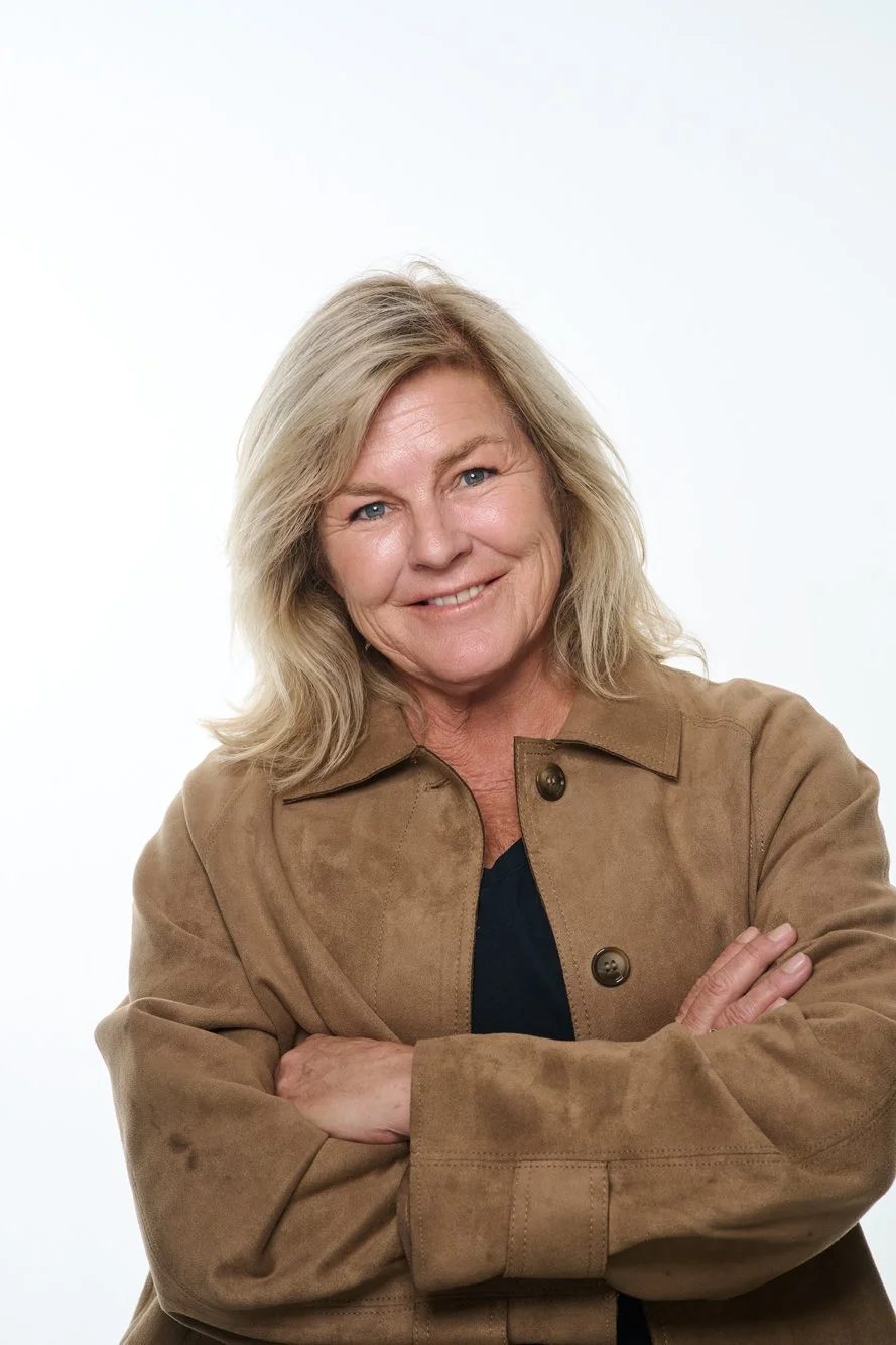 A middle-aged woman with blonde hair smiling, wearing a light brown jacket and crossing her arms, against a white background.