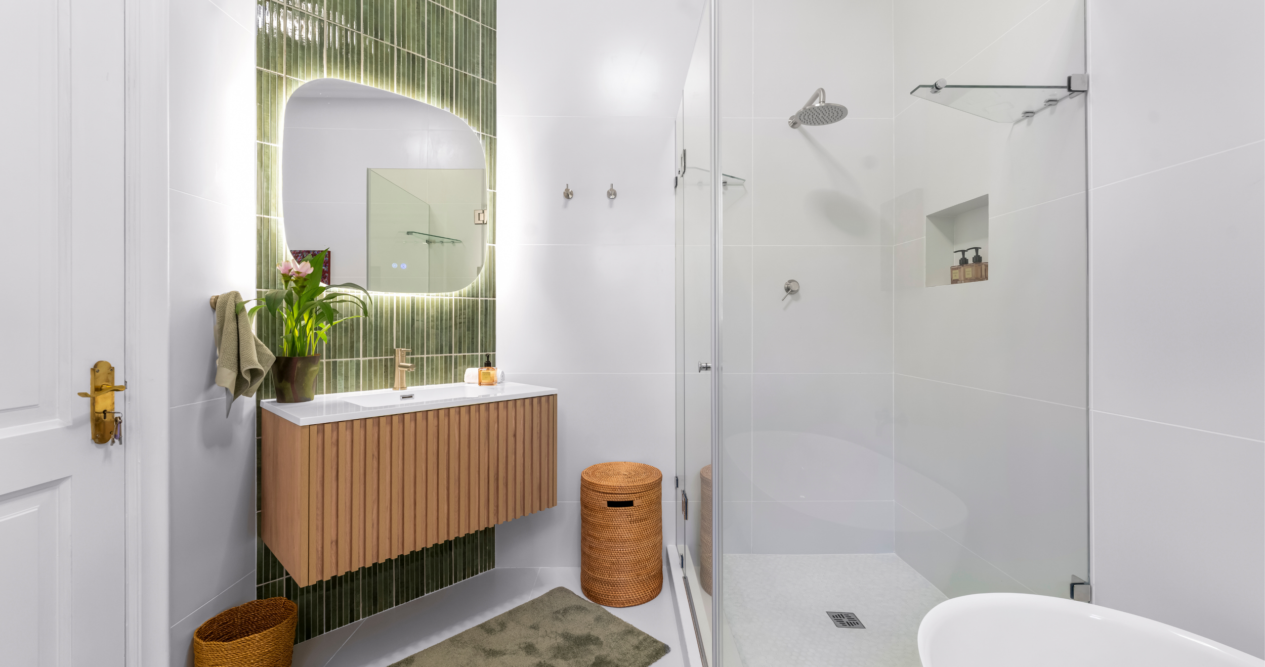 Modern bathroom with a green tiled wall, wooden vanity, mirror, potted plant, shower with glass door, and minimalist decor.