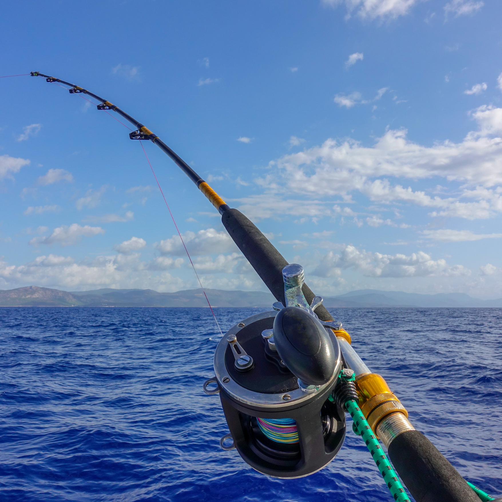 Close-up of a fishing rod and reel set against a blue ocean and sky with some clouds.