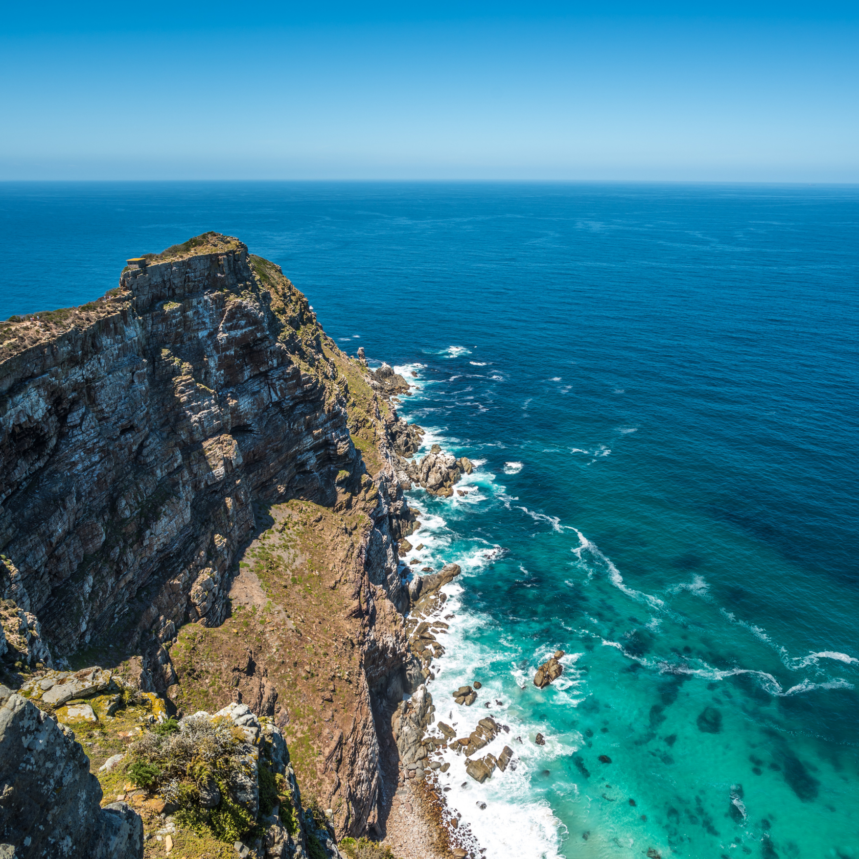 A rugged cliff extending into the ocean under a clear blue sky, with waves crashing at the base of the cliff.
