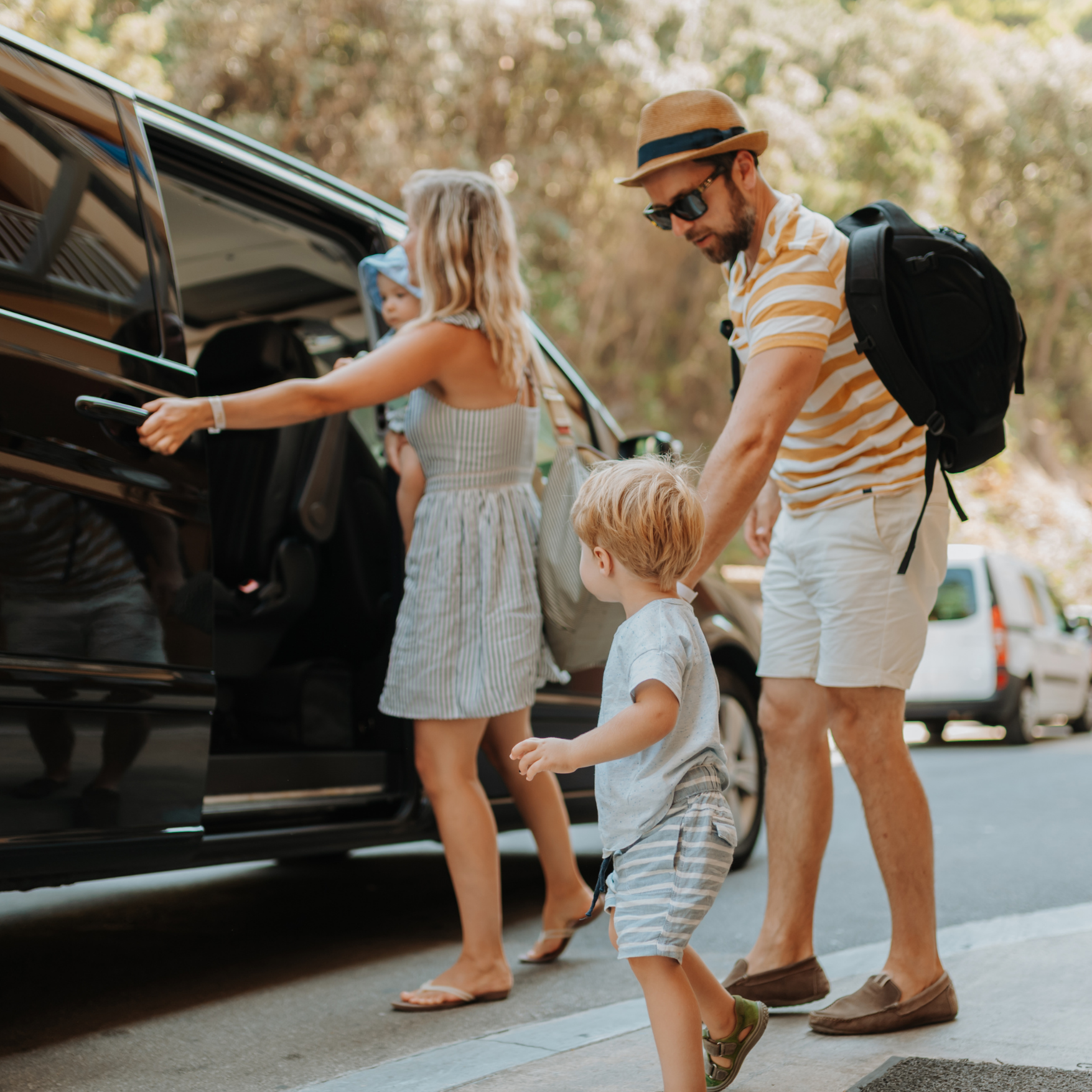 A family of four with children and an adult man unloading luggage from a black car, standing outdoors in a park with trees in the background.