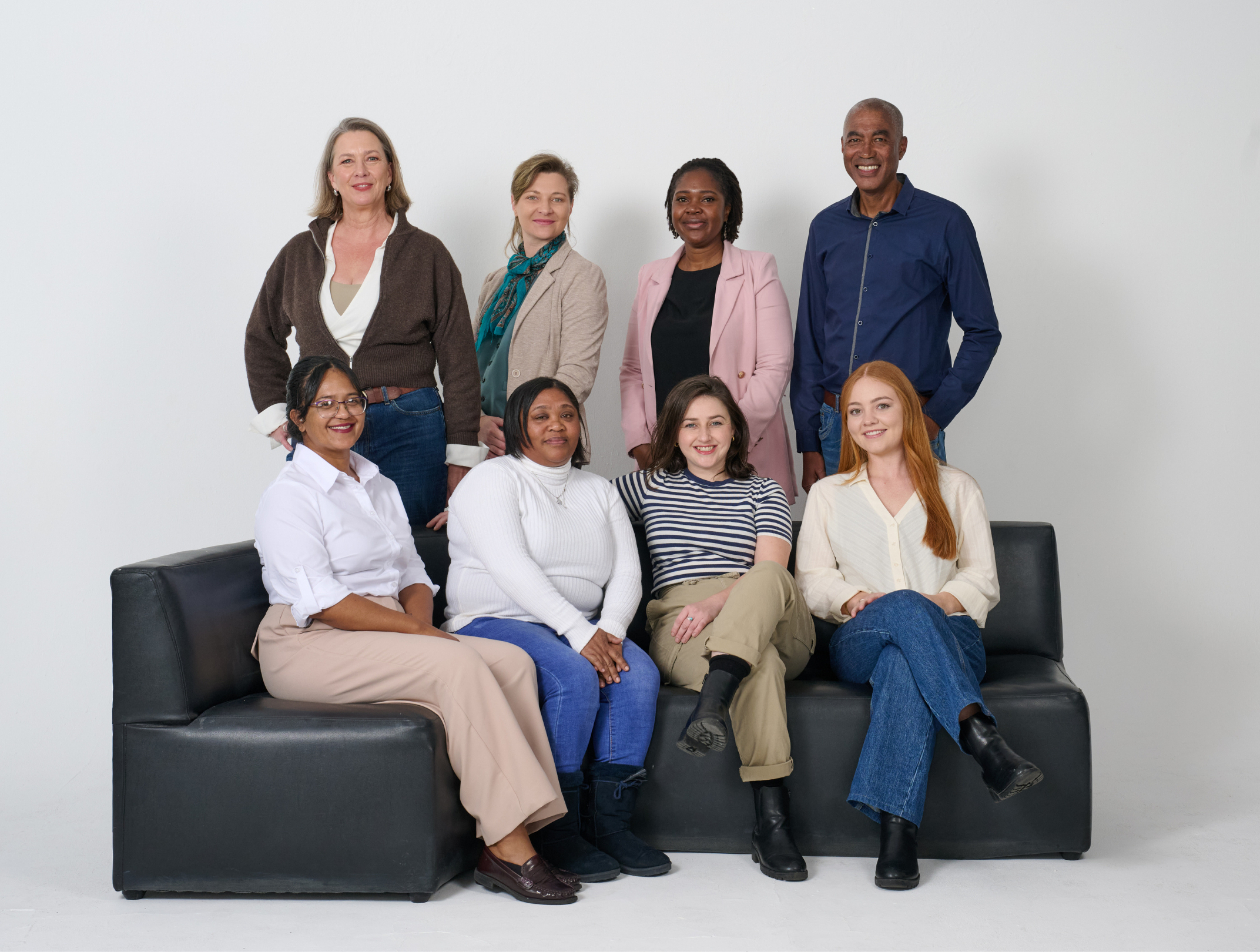 Group of diverse professional women with light and dark skin tones, some sitting on a black sofa and others standing behind, posing for a photo against a plain white background. They are dressed in business casual attire and smiling.