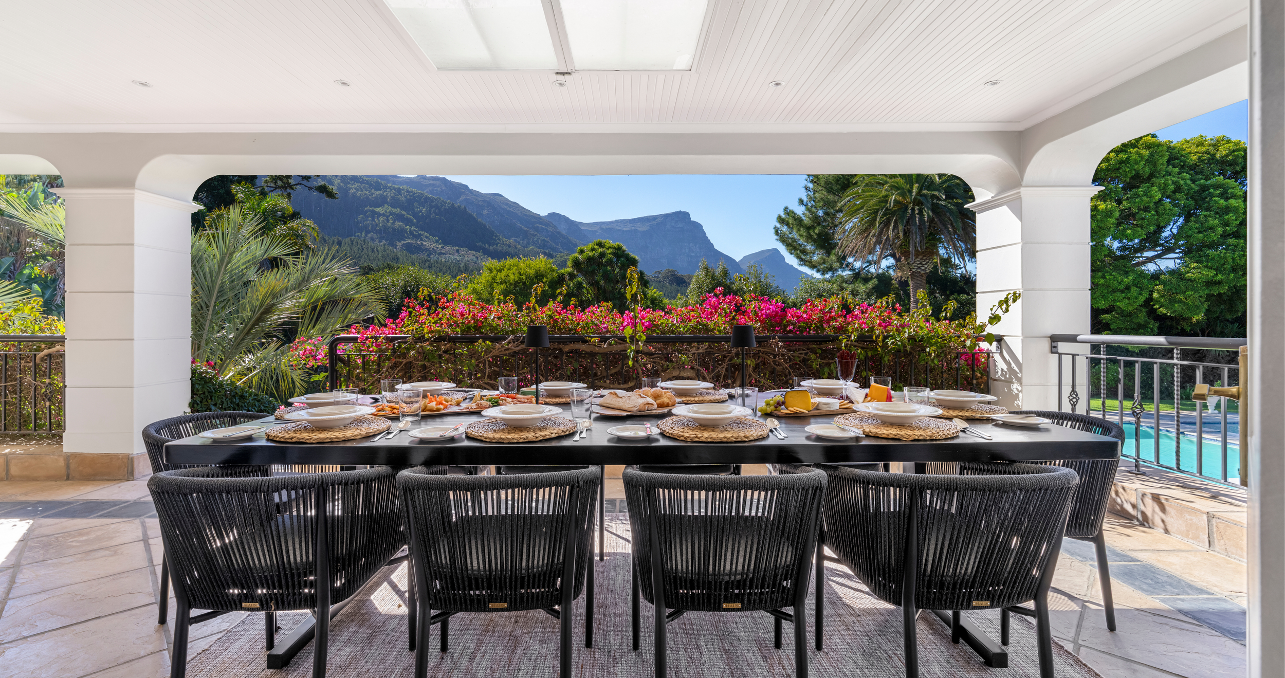 Outdoor dining table set with plates, glasses, and food on a patio, overlooking mountains and lush greenery.
