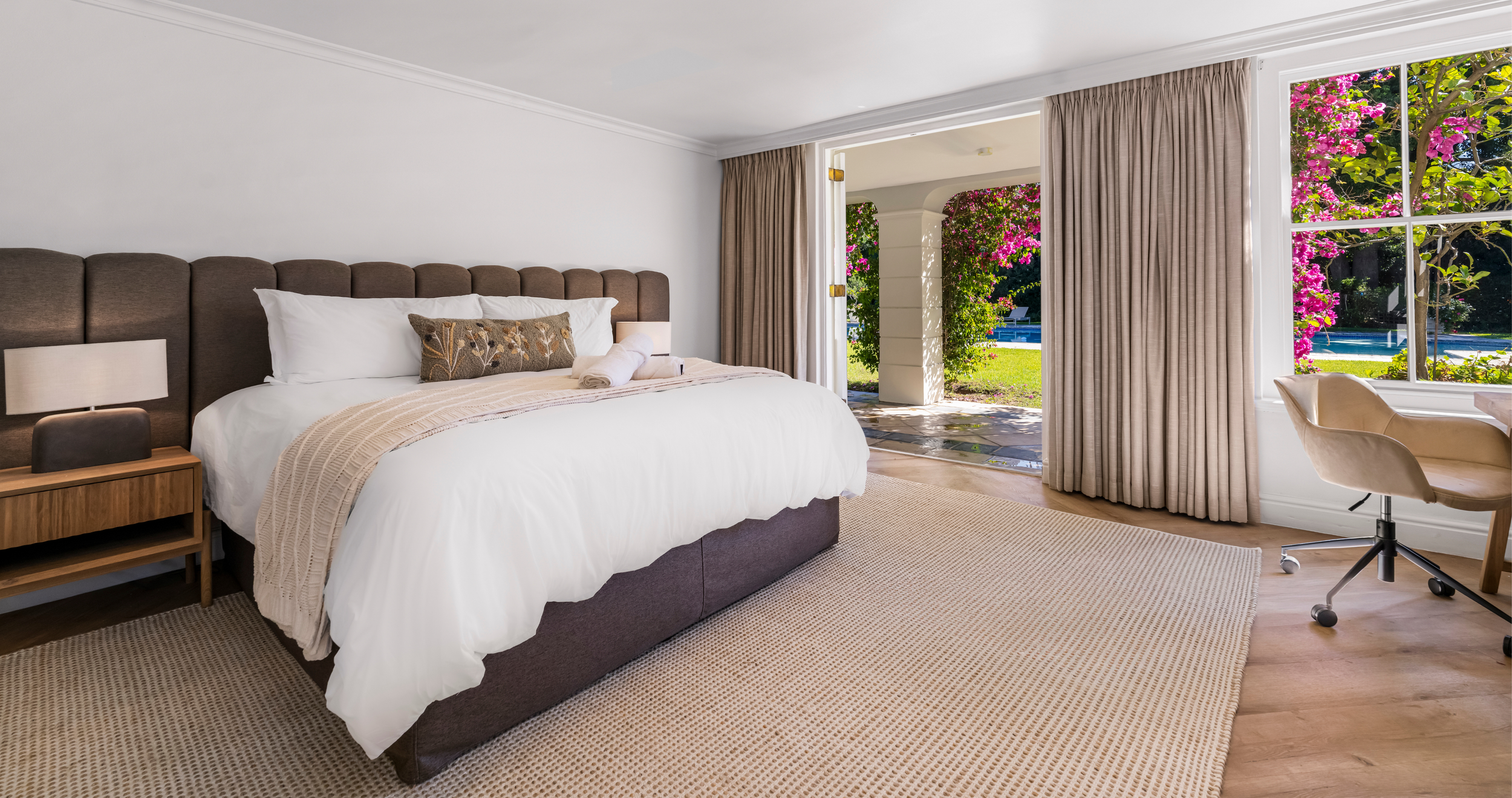 A bedroom with a large bed, bedside table, lamps, and a window with beige curtains. Outside the window, bushes with pink flowers and a fountain are visible.