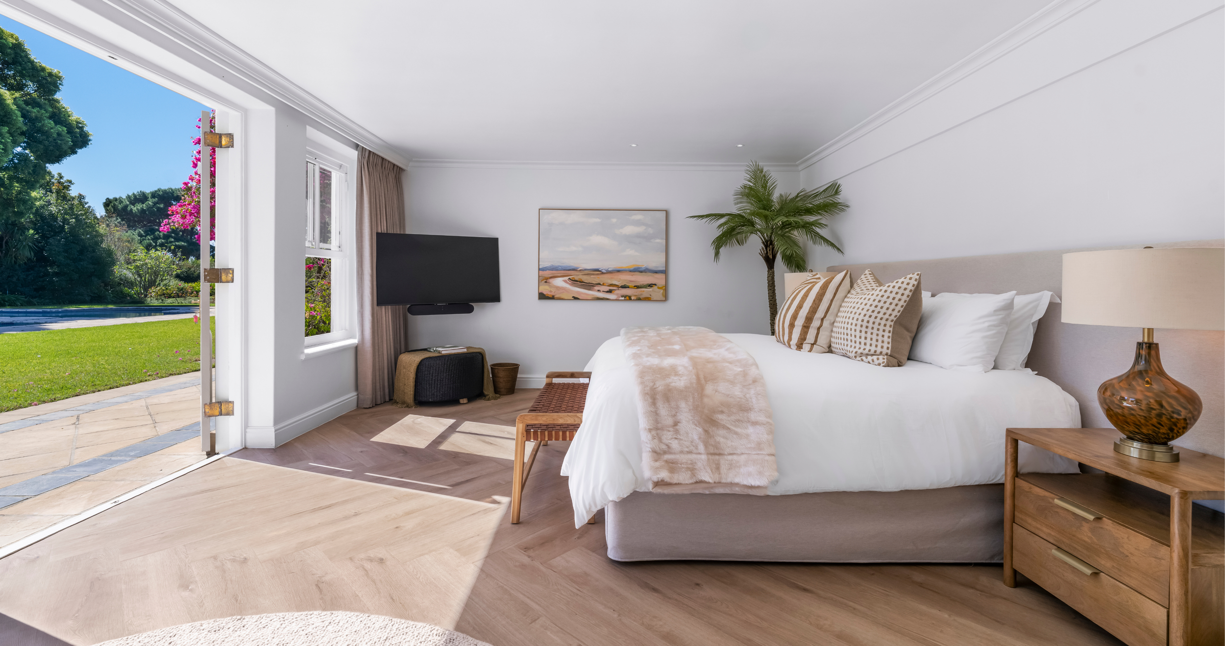 Bright bedroom with a large bed, pillows, a blanket, a nightstand with a lamp, a TV, artwork, and a view outside through open doors and windows showing greenery and a blue sky.