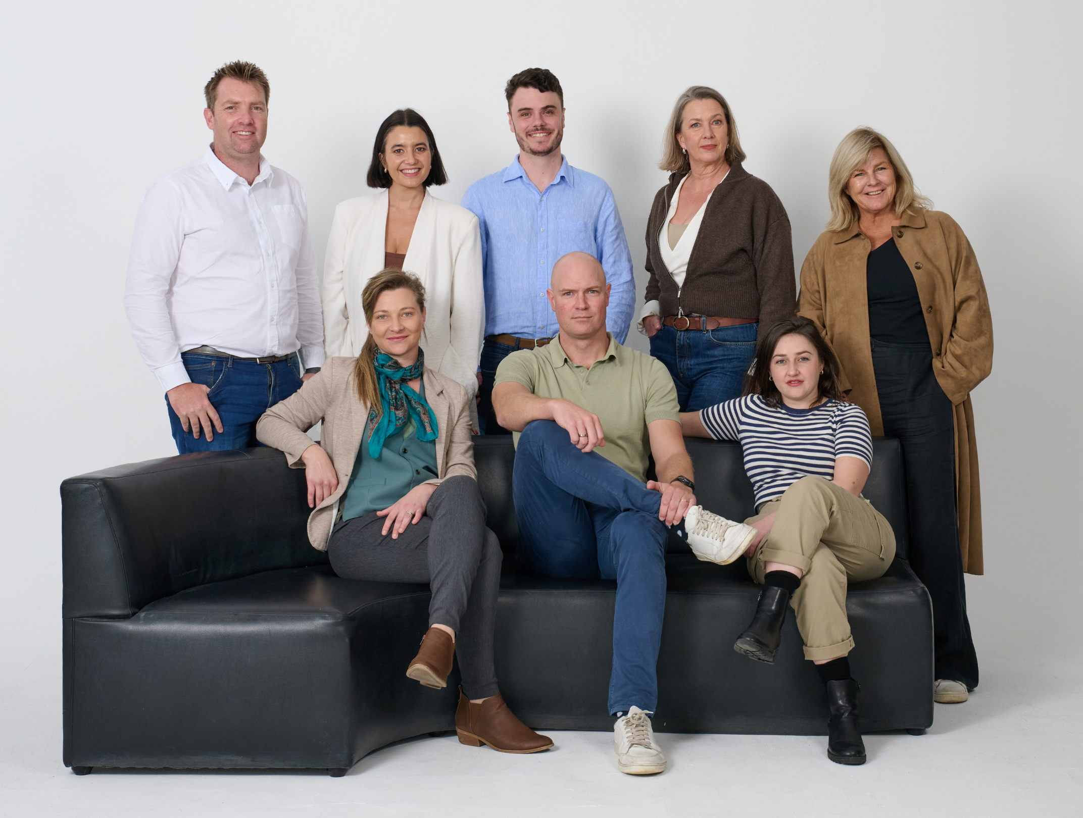 A diverse group of nine people, including men and women of various ages, posing together against a plain white background. Some are standing while others are seated on a black leather couch, smiling for the camera.
