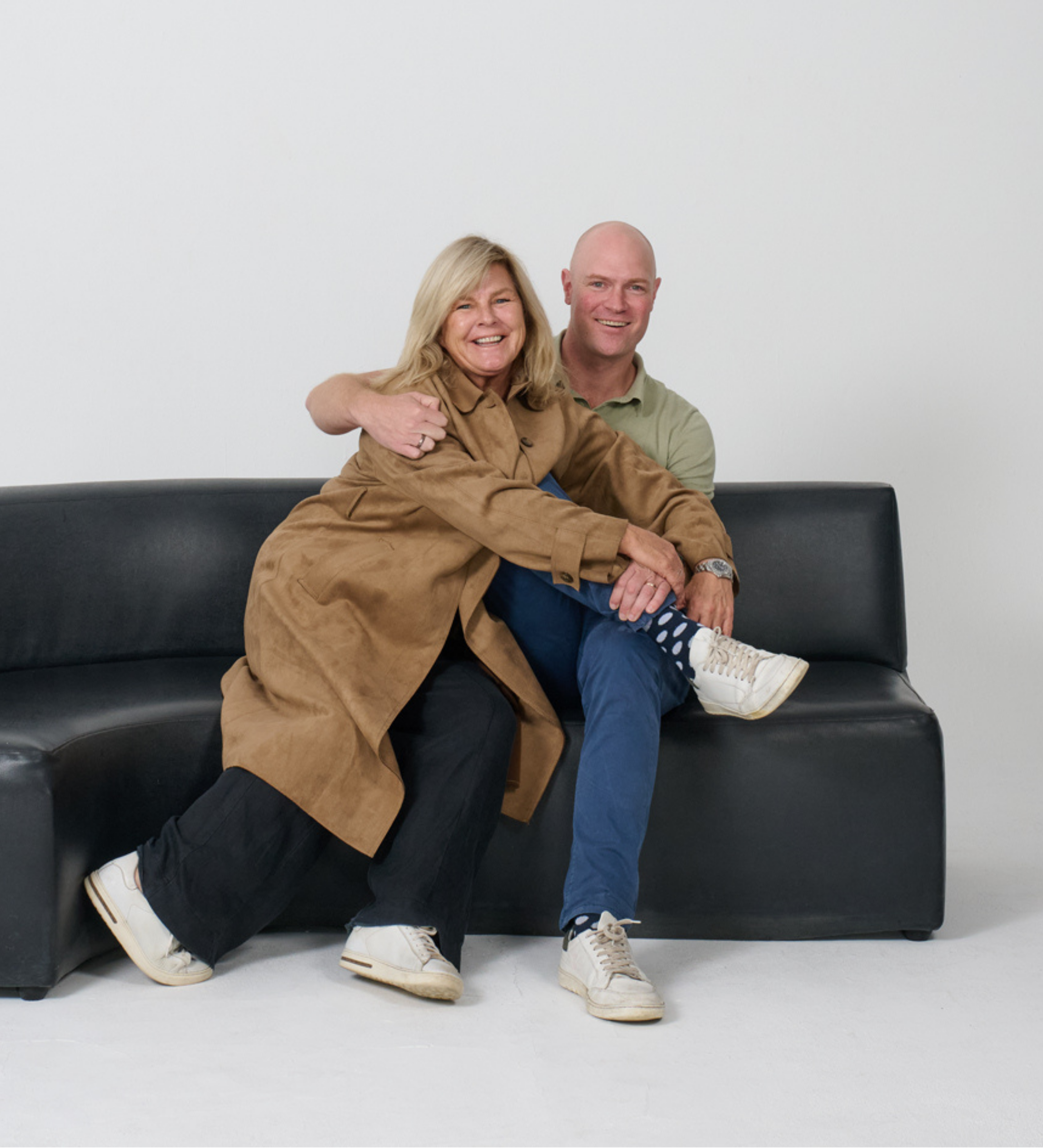 A smiling woman with shoulder-length blonde hair and a man with a shaved head sitting together on a black sofa against a white wall, both dressed casually in beige and light green shirts, with the woman's arm around the man's shoulders.