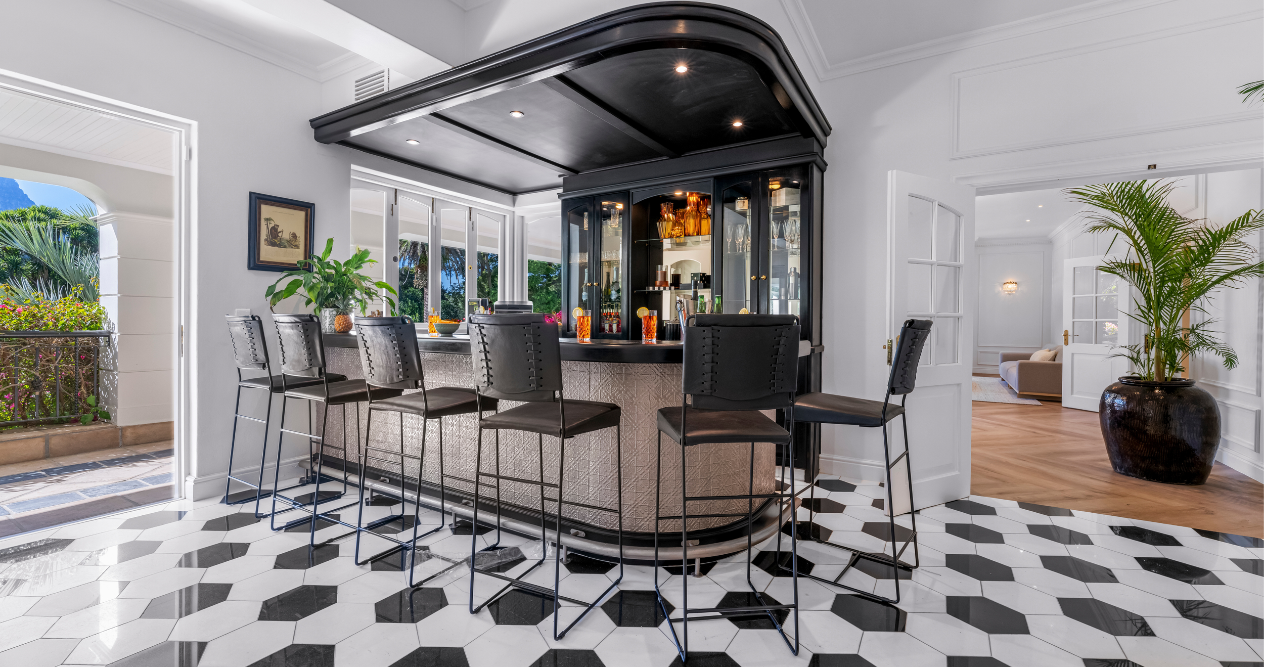 Interior view of a stylish home bar with black cabinetry, bar stools, large windows, and decorative plants in a well-lit room.