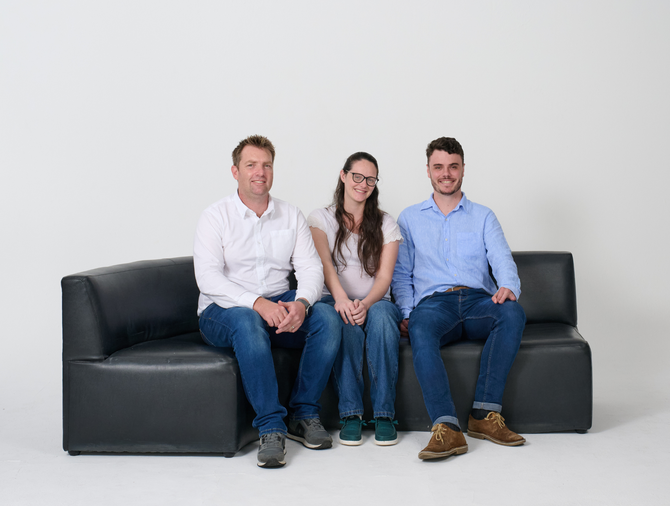 Three people sitting on a black couch against a plain white wall, smiling at the camera, with the man on the left, a woman in the middle, and a man on the right.