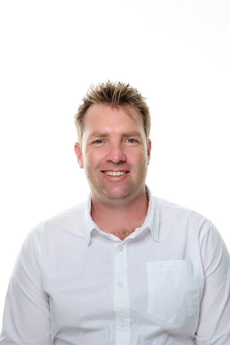 Portrait of a man with short, spiked blonde hair, wearing a white button-up shirt, smiling against a plain white background.