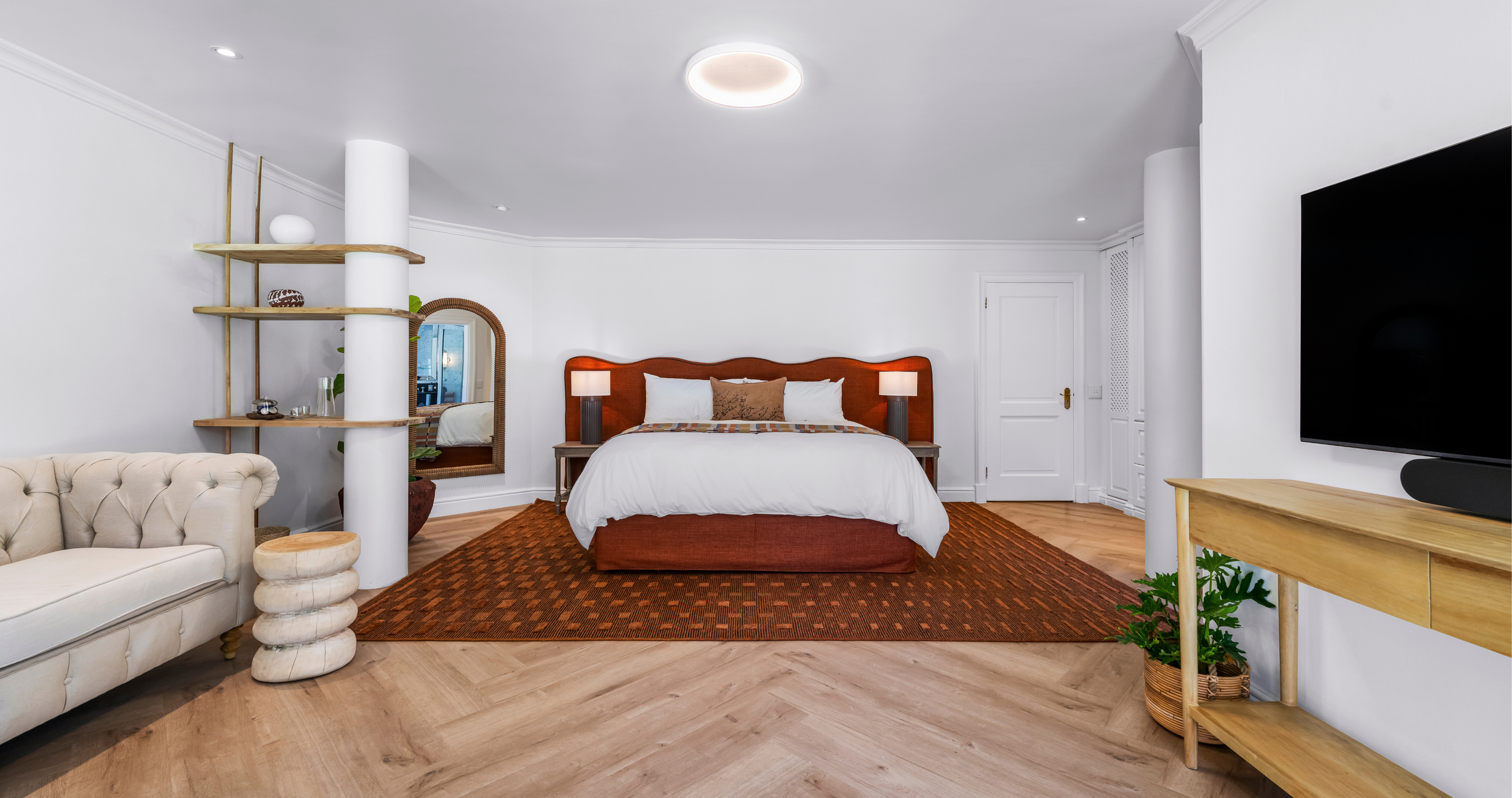 A bedroom with a large bed with a wooden headboard, two nightstands with lamps, a mirror, a white sofa, and a TV on a wooden stand. The room has white walls, a red patterned rug, and wooden flooring.