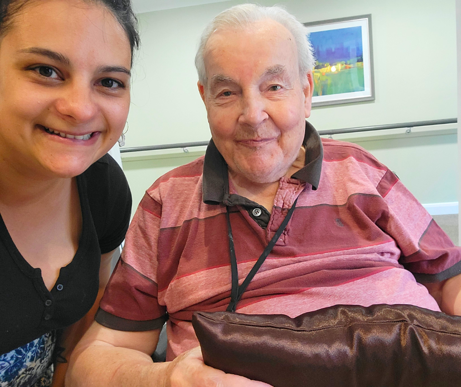 A young woman and an elderly man smiling for a selfie photo indoors, with a painting on the wall behind them.