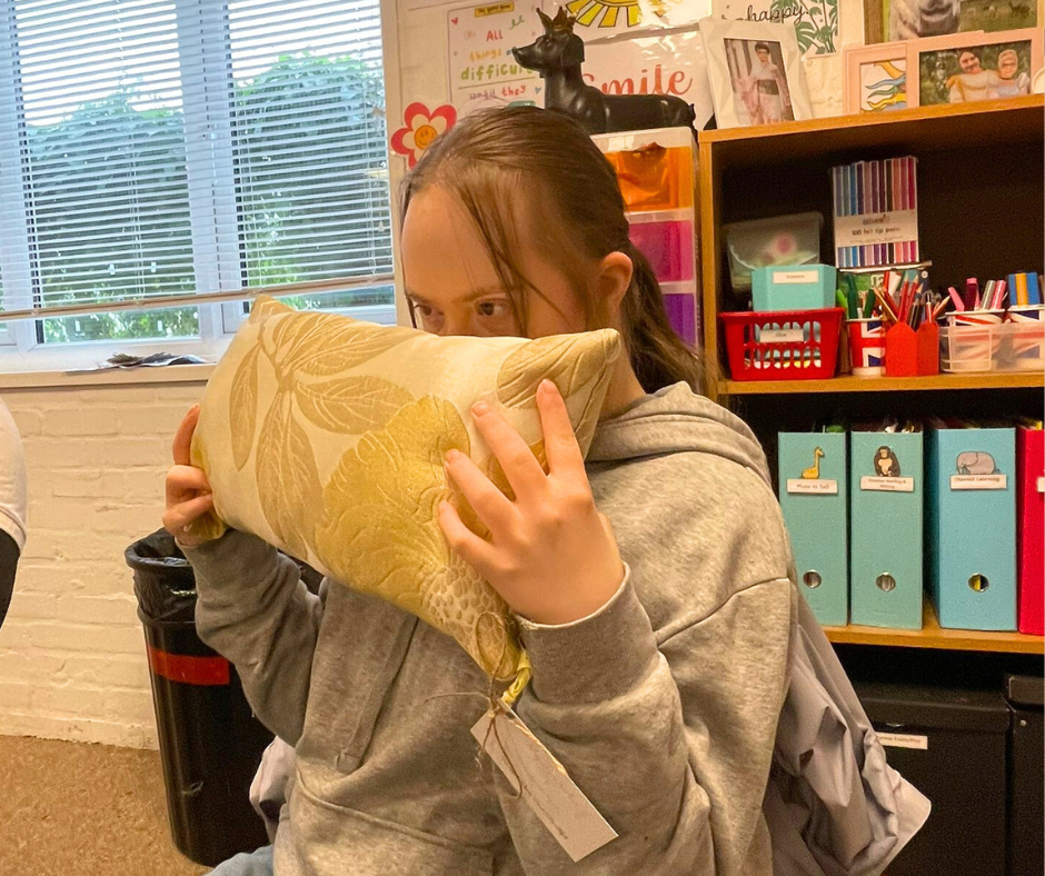 A girl sitting in a classroom holding a decorative pillow up to her face. She is wearing a gray hoodie and has a backpack. The background shows shelves with colored storage bins, books, and classroom decorations.