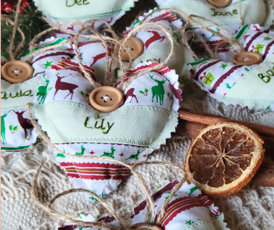 Close-up of fabric heart ornaments with Christmas reindeer and gifts patterns, featuring buttons, twine bows, and personalized names like "Lily". Dried citrus slice and wooden stick add festive decor.