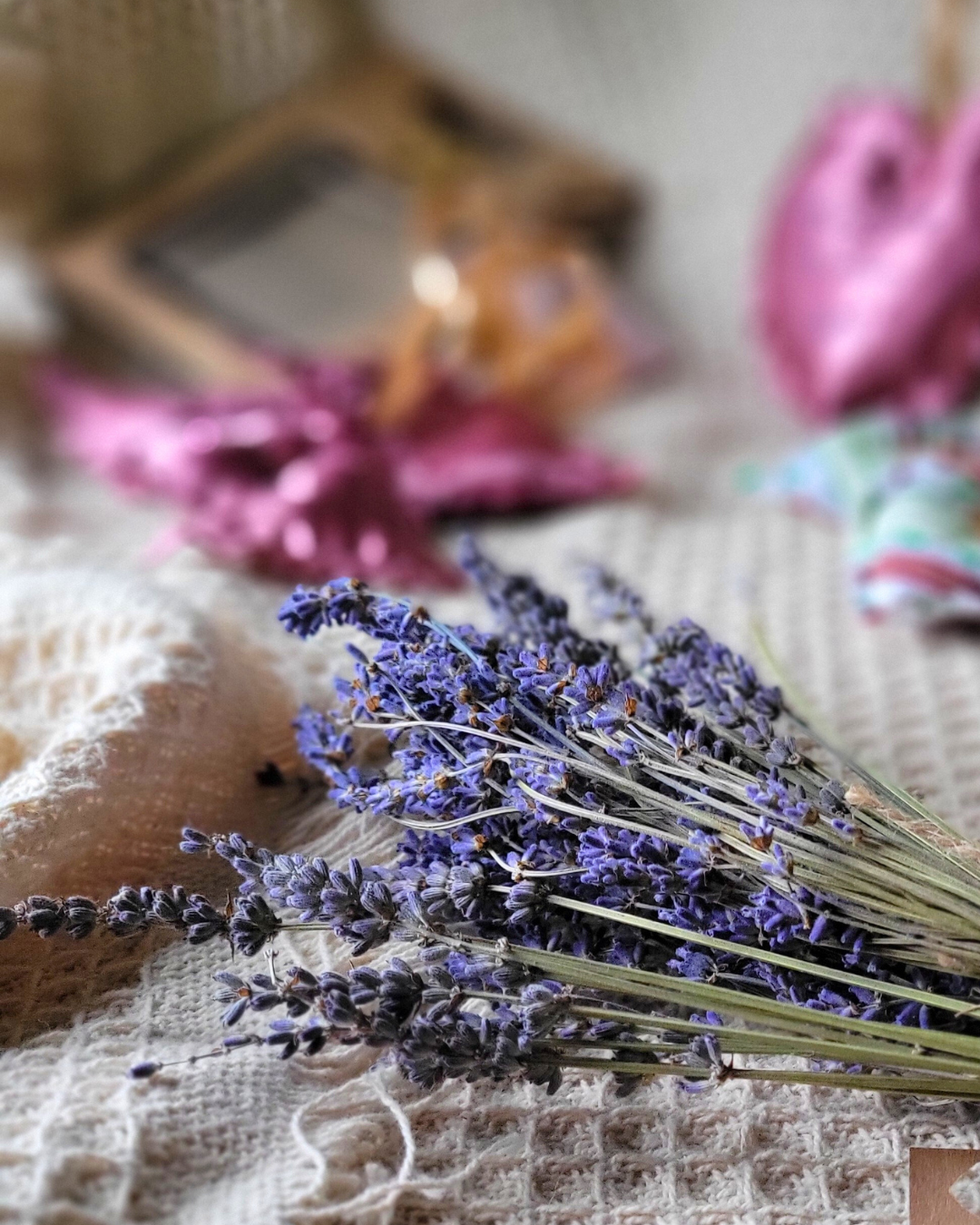 A bunch of dried lavender flowers lying on a textured cream-colored surface with a blurred background of a mirror, pink paper flowers, and colorful cloth.