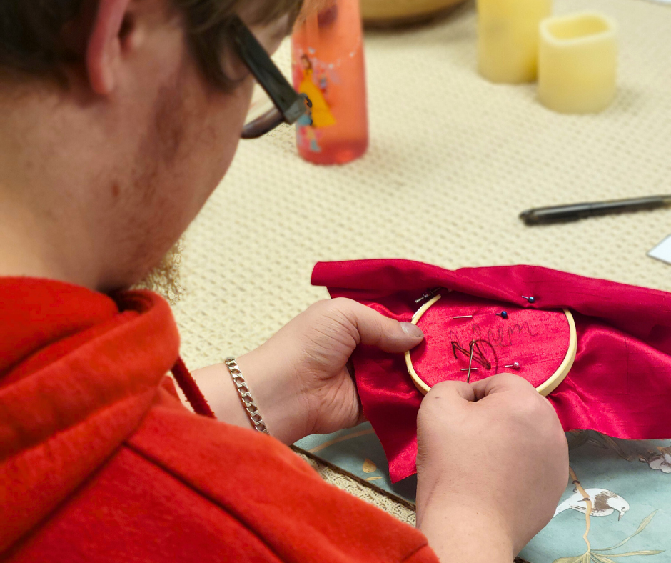 A person working on a sewing project with an embroidery hoop, red fabric, and sewing pins, on a table with craft supplies.