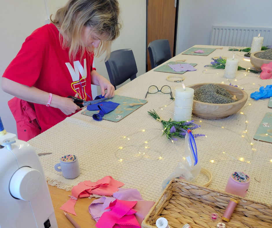 Woman in red shirt cutting fabric at a crafting table decorated with candles, flowers, and crafting supplies for a DIY project.
