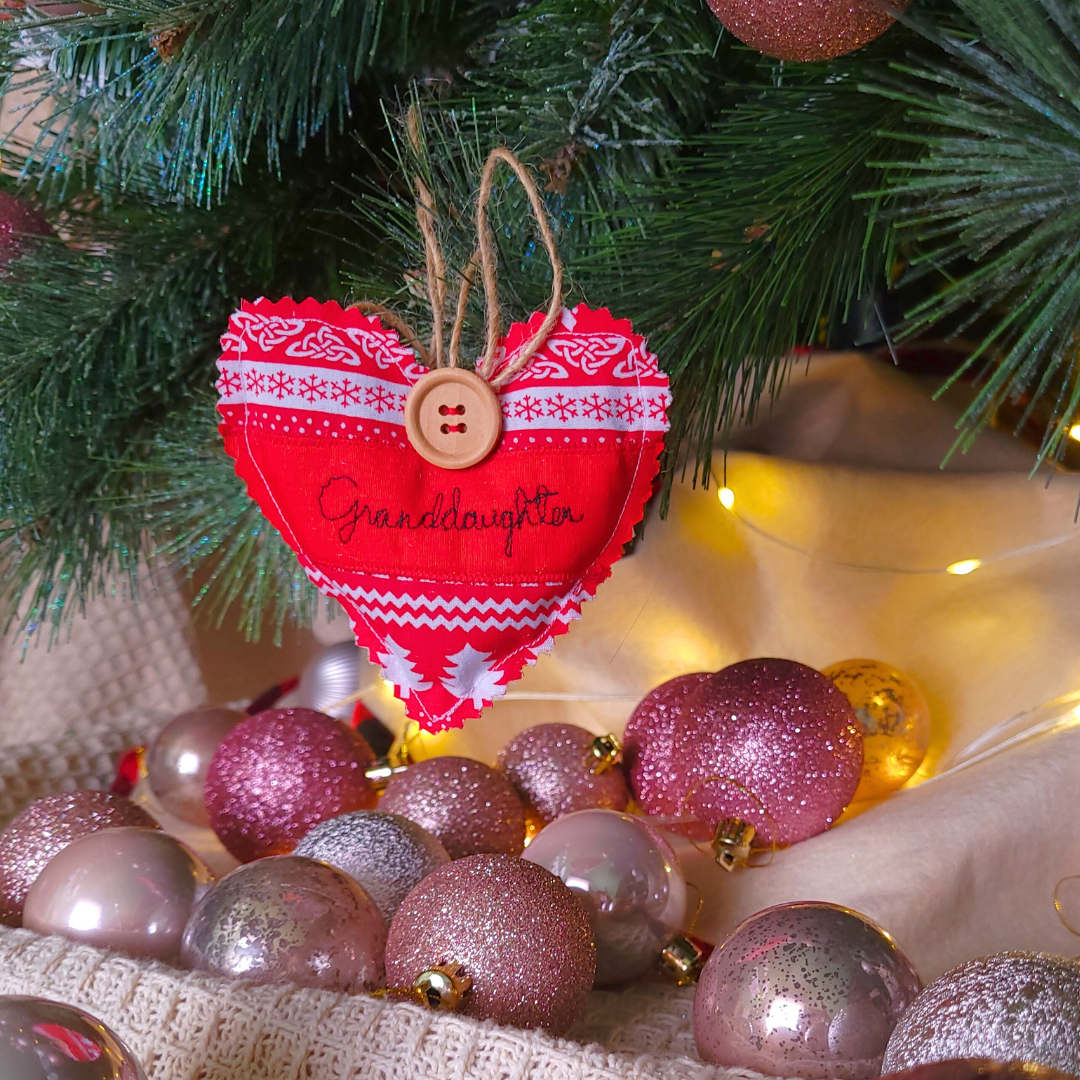 Decorated Christmas tree branch with pink and gold ornaments, a heart-shaped embroidered Christmas decoration saying 'Granddaughter', and warm yellow lights.