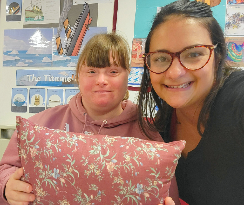 A woman with glasses smiling next to a girl holding a pillow with a floral pattern. The girl is in a classroom with posters about the Titanic and other educational materials on the wall.