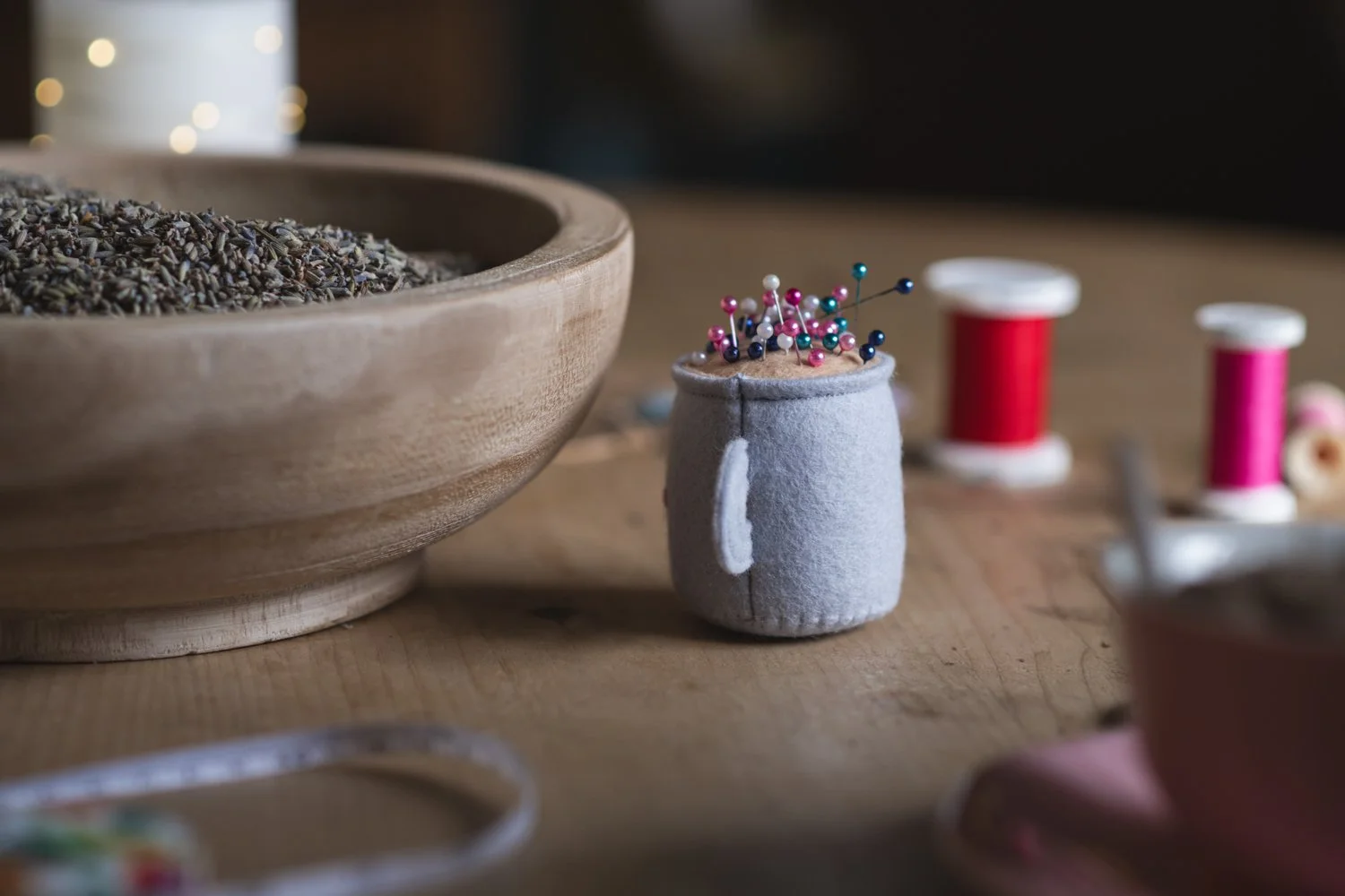 A small fabric pin cushion with multicolored sewing pins, next to a wooden bowl filled with dried lavender on a wooden surface, with spools of red and pink thread in the background.