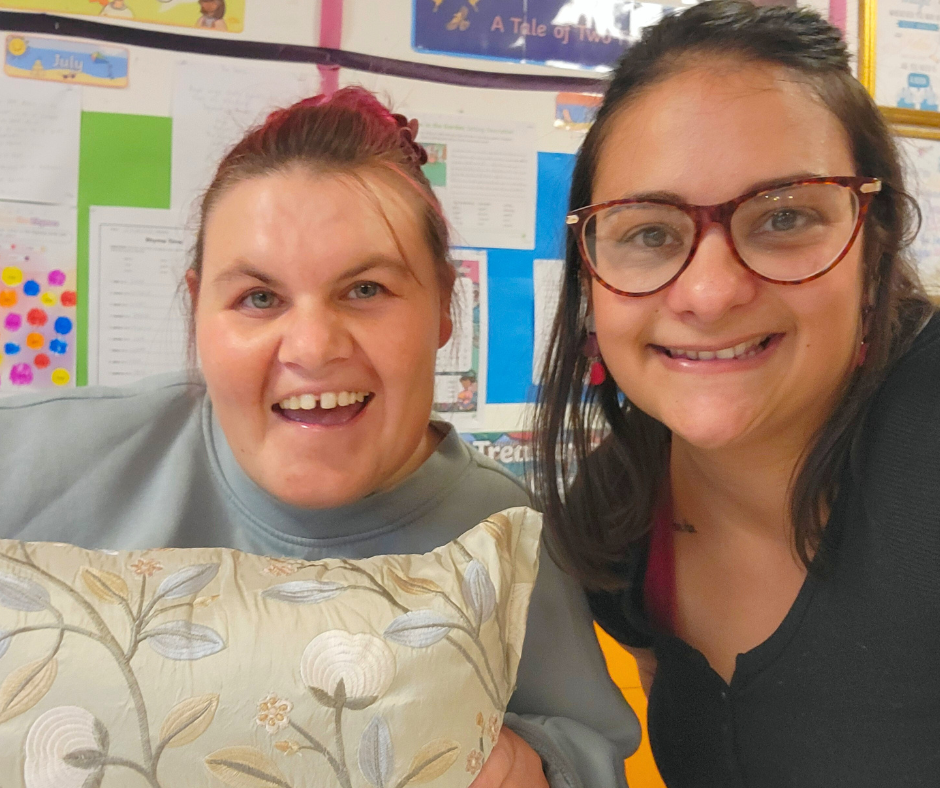 Two smiling women taking a selfie in a classroom with colorful posters in the background. One woman is holding a decorative pillow with a floral pattern.