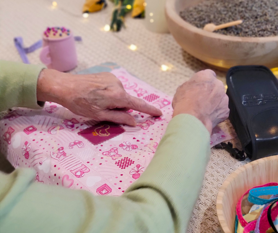 Elderly person's hands holding pink fabric while sat at a table, surrounded by craft supplies and lavender seeds in a bowl.