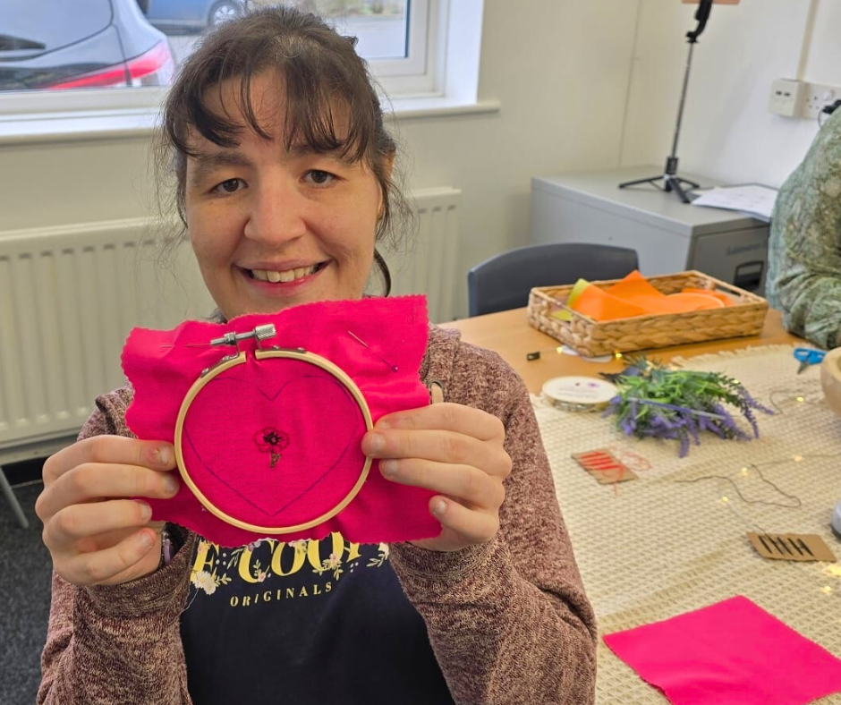 A woman smiling and holding an embroidery hoop with pink fabric and embroidered heart design in a craft room.