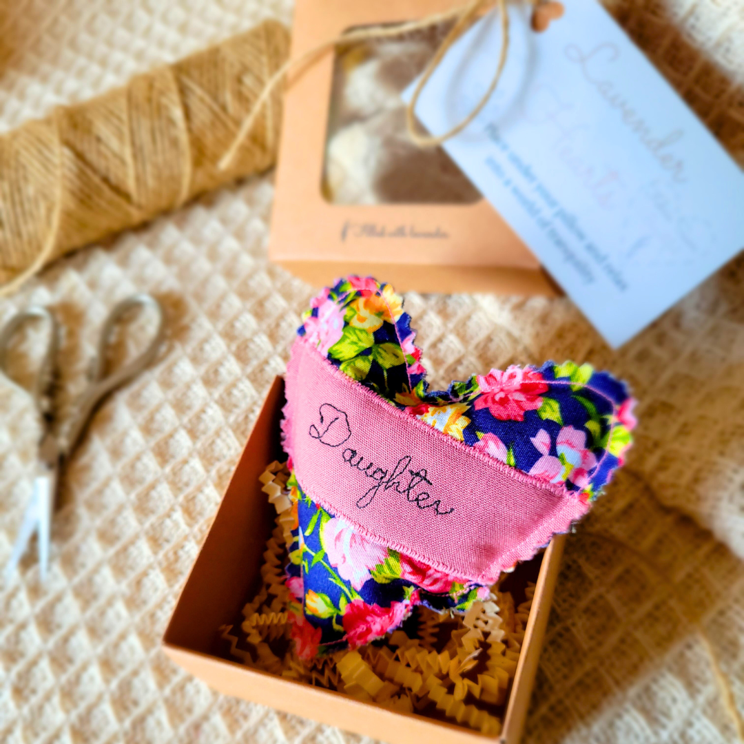 A small fabric heart with a floral pattern and pink label with 'Daughter' embroidered on it, placed inside a pink box filled with yellow crinkle paper. In the background, there are beige scissors, a bamboo whisk, and a partially visible box with a clear window and a blue note attached.
