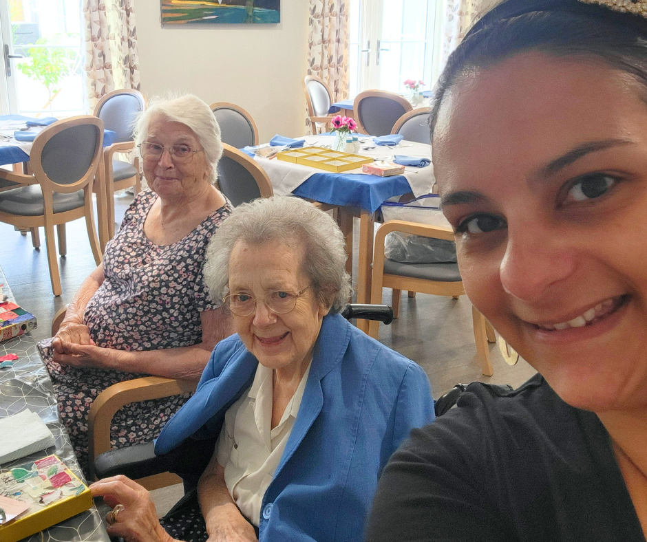A young woman taking a selfie with two elderly women seated at a workshop table in a care home.