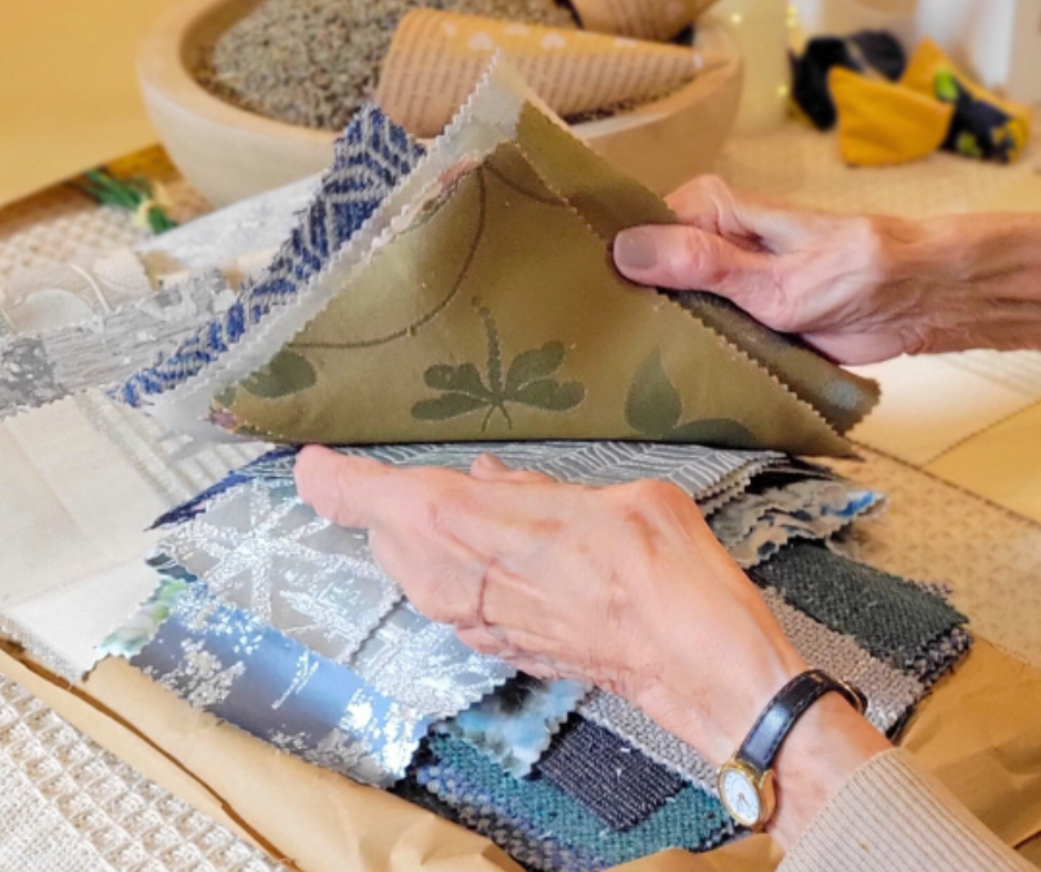 Hands of an elderly woman choosing a variety of fabrics in a pile on a table with cream tablecloth and a wooden bowl of lavender seeds