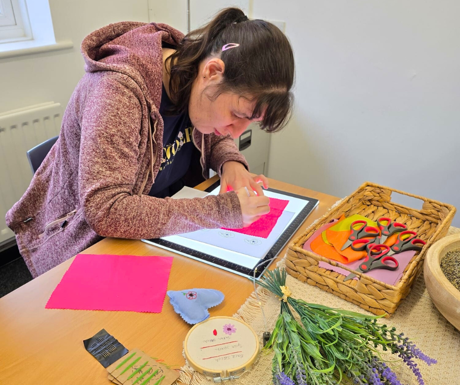 A woman using a drawing tablet on a table, surrounded by craft supplies including fabric, scissors, and greenery, in a well-lit room.