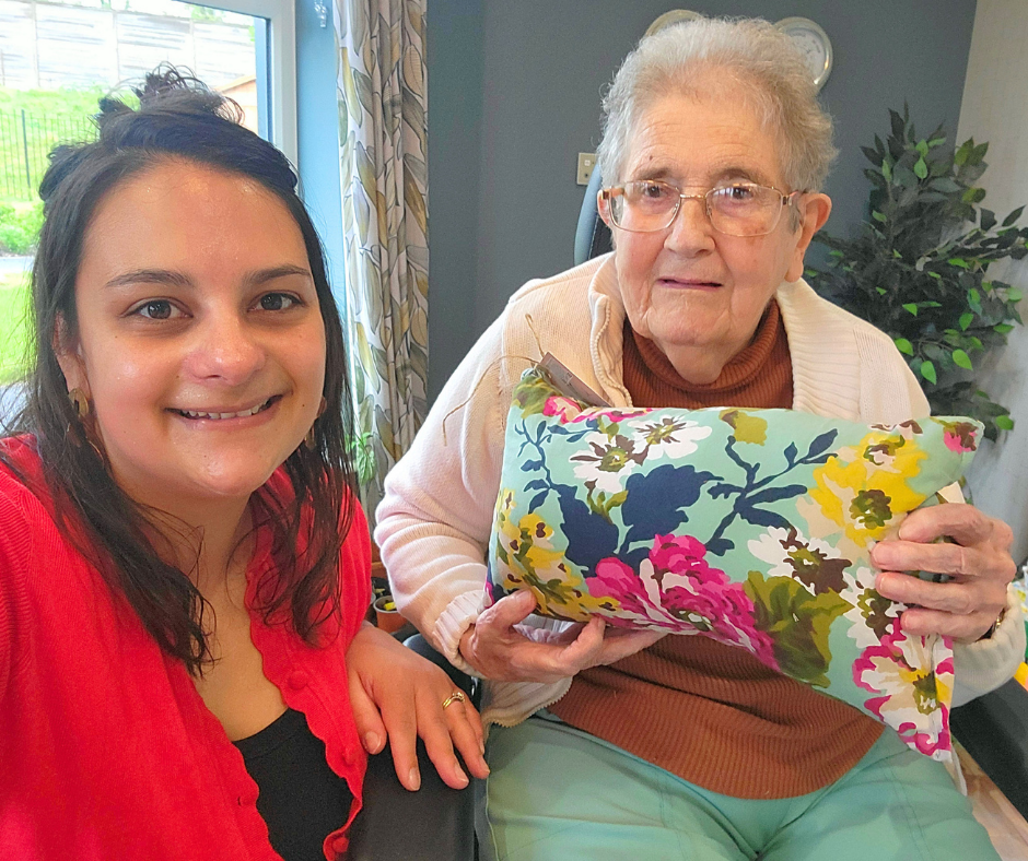 Young lady with long dark hair and red top is smiling and kneeling next to an elderly women sat in a chair holding a green flowery cushion
