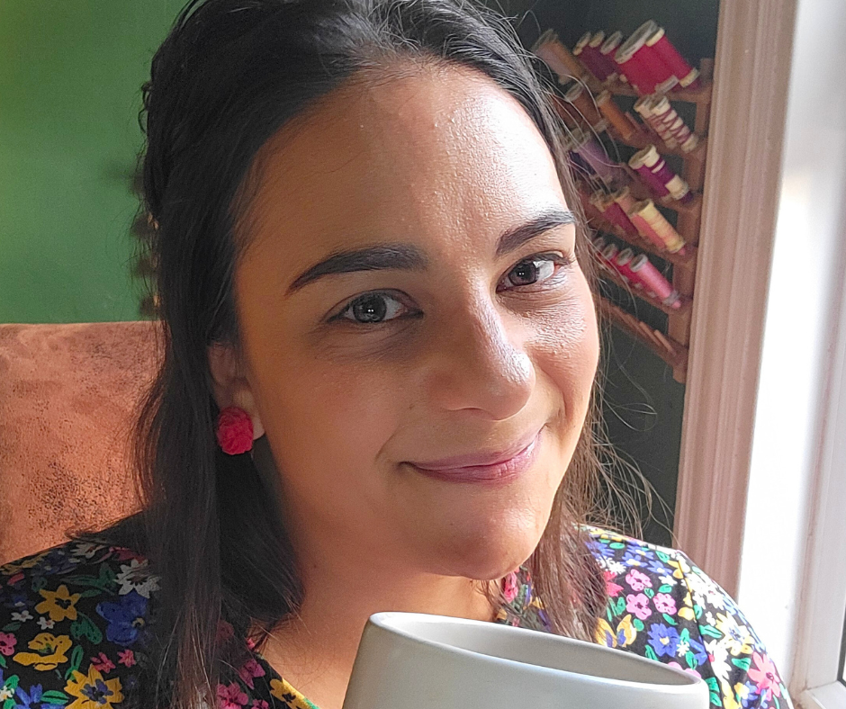 A woman with dark hair and pink earrings holding a mug, smiling, with shelves of cotton reels in the background.