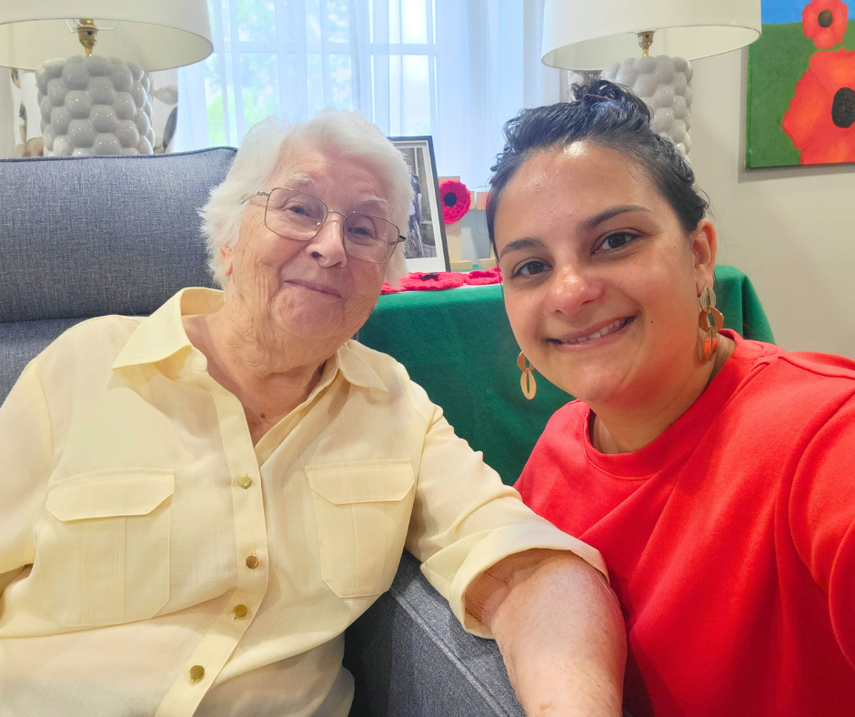 Young women with dark hair tied up wearing red jumper is sat next to an elderly women with grey hair wearing yellow blouse, both smiling