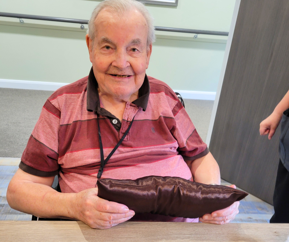 Elderly man with pink and red top is sat at a table smiling holding a brown cushion