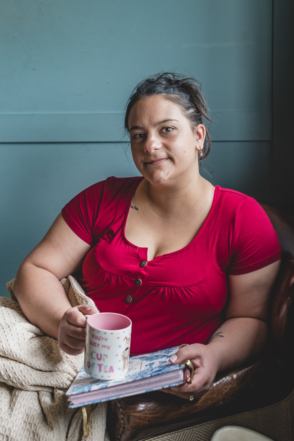 A woman in a red shirt sitting indoors holding a colorful mug and a notebook.