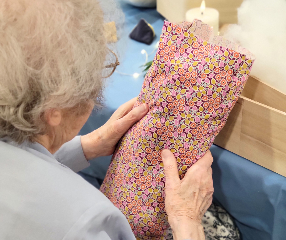 Image shows the back of an elderly woman's grey hair, over her should shows she is holding a pink flowery cushion