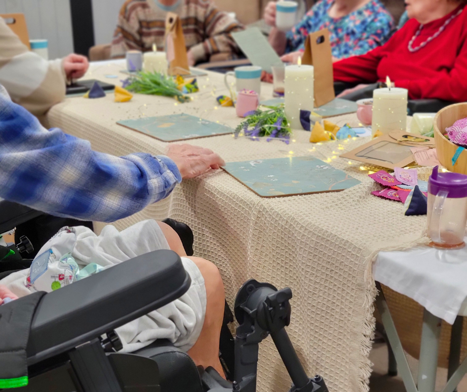 A long narrow table with cream tablecloth is surrounded by elderly residents, one in a black wheelchair. The table has green placemats, mugs, artificial lavender and candles
