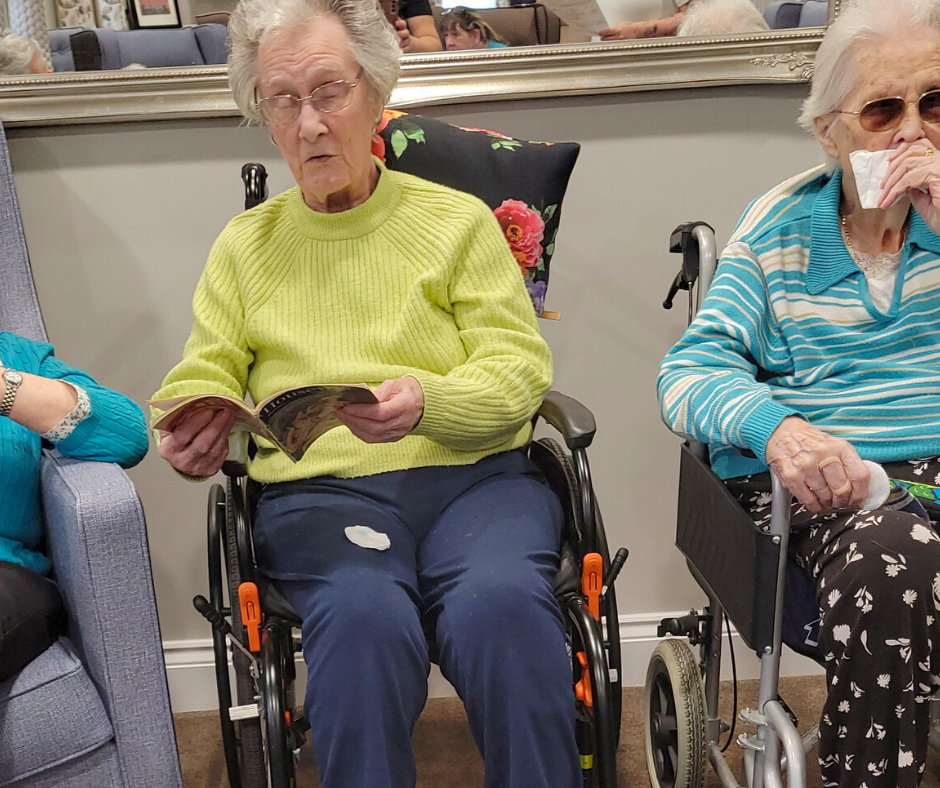 Elderly woman with green jumper and navy trousers sat in a black wheelchair looking at an old book