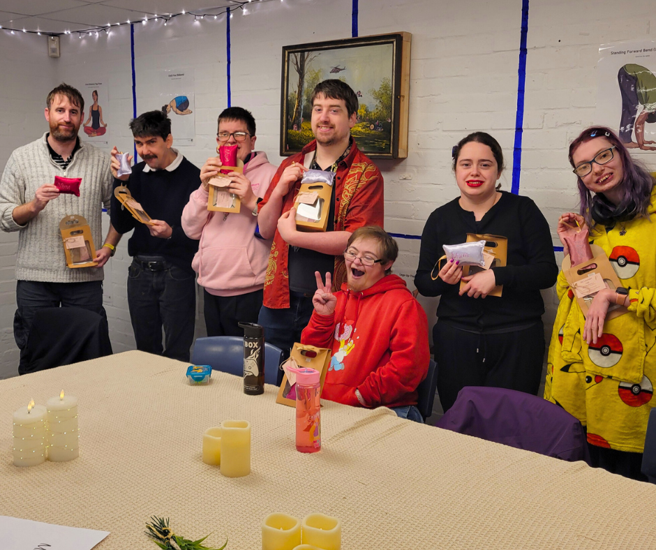 Group of seven people standing behind a table, holding colorful socks packaged in boxes, in a decorated room with candles on the table, paintings on the wall, and string lights overhead.