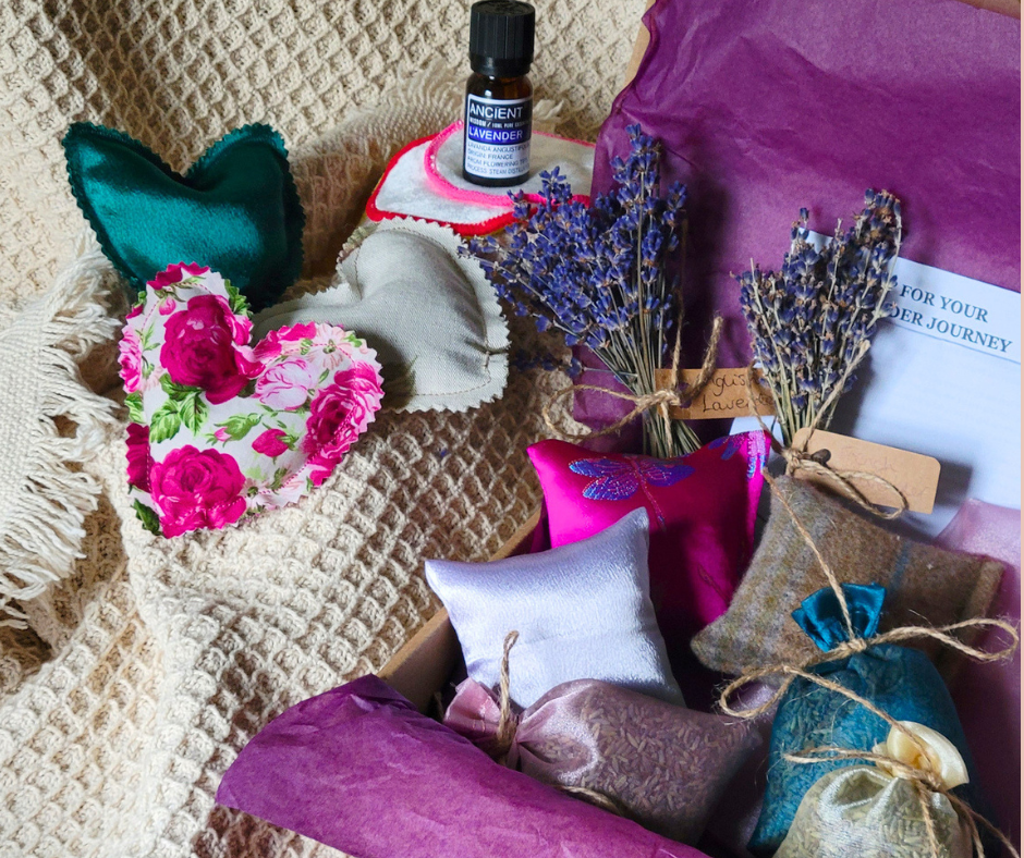 Assorted small gift boxes wrapped in colorful fabric and tied with twine, lavender bundles, a bottle of lavender essential oil, a pink and white floral heart-shaped fabric item, and a purple paper bag.