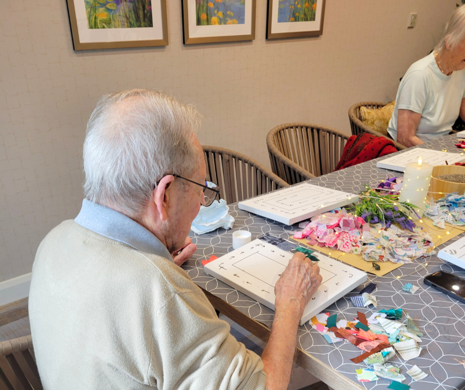 An elderly man working on a craft project at a decorated table with torn paper, flowers, and other craft supplies, while another elderly woman is in the background, with framed artwork on the wall behind.
