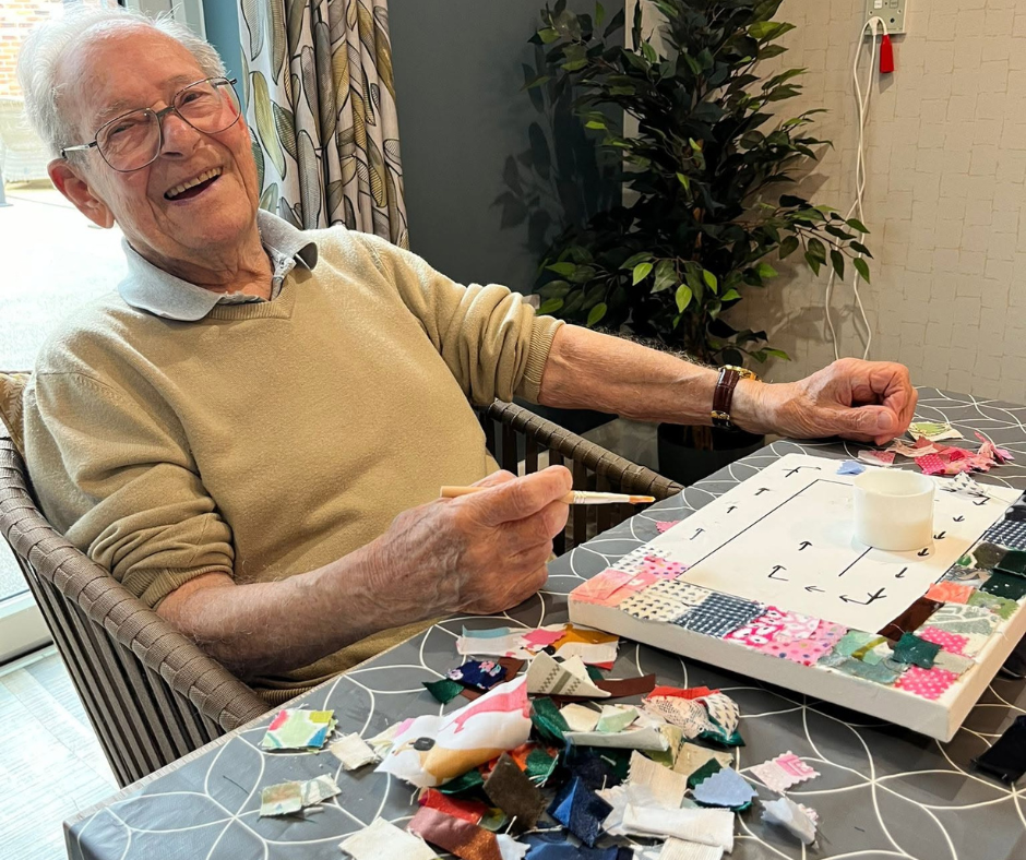 An elderly man with glasses, wearing a beige sweater, sitting at a table and smiling participating in a fabric activity. The table has various fabric scraps. There is a plant and curtains in the background.