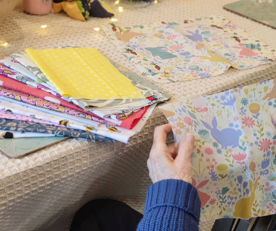 A person in a blue sweater folding colorful fabric with bunny and floral patterns on a table with other fabric pieces and crafting supplies.