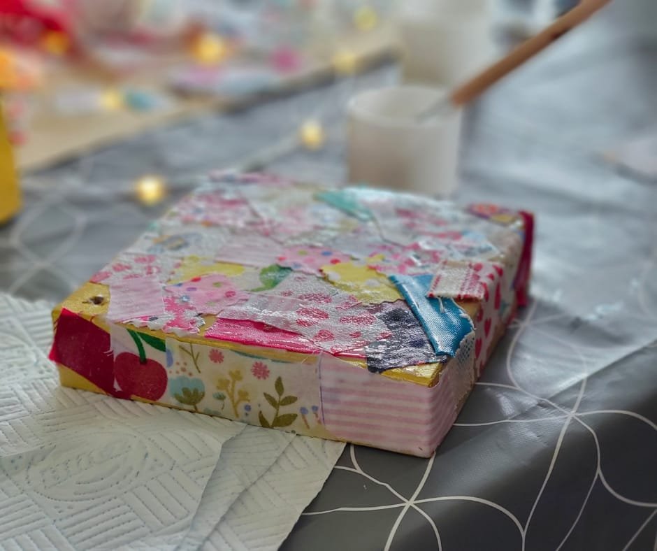 A cardboard box decorated with colorful, floral-patterned tape, placed on a table with a paper towel nearby. In the background, there is a white cup and a hand using a paintbrush, and blurred craft supplies.