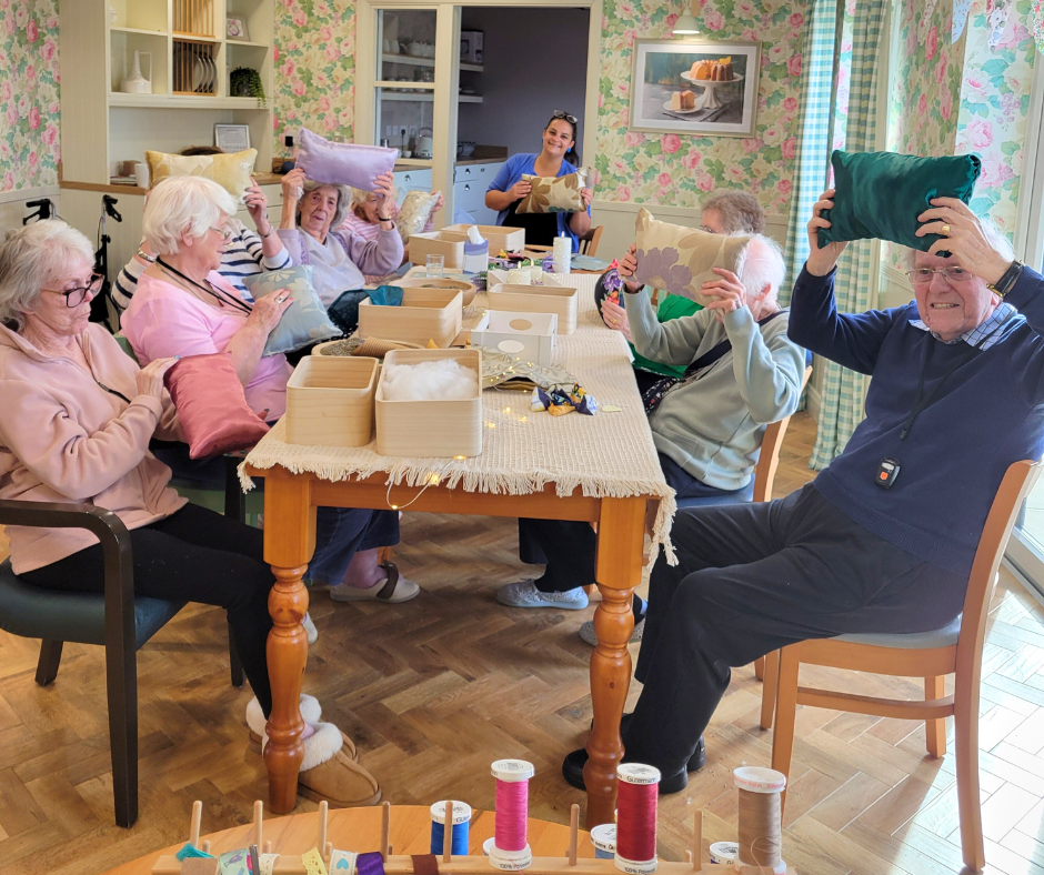 Group of elderly care home residents sat at a rectangular table holding up their cushions made while participating in a workshop with Fabric Sewn Beautiful.