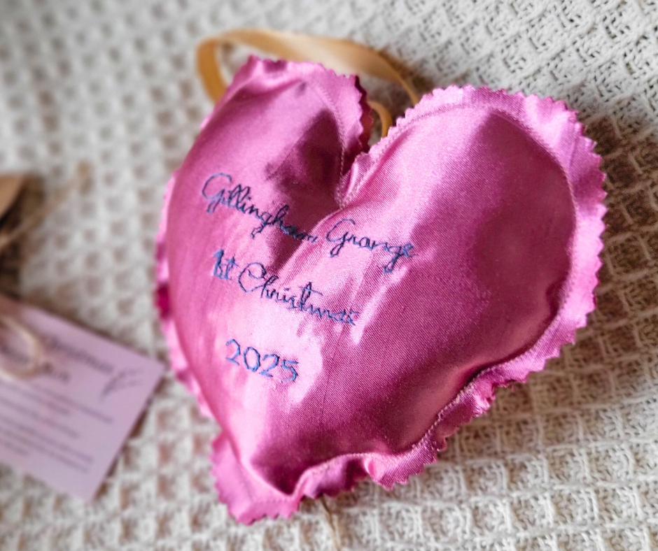Pink satin heart-shaped sachet with embroidered message for a family Christmas tree, reading "Gillingham Grange, Christmas 2025" placed on a textured surface.